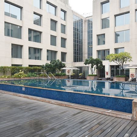 Holiday Inn New Delhi Aerocity By Ihg Exteriér fotografie The photo shows a modern outdoor swimming pool situated in a courtyard surrounded by several buildings. The pool has a sleek design with a blue tiled edge and is bordered by wooden decking. There are a few trees and plants around the area, adding som