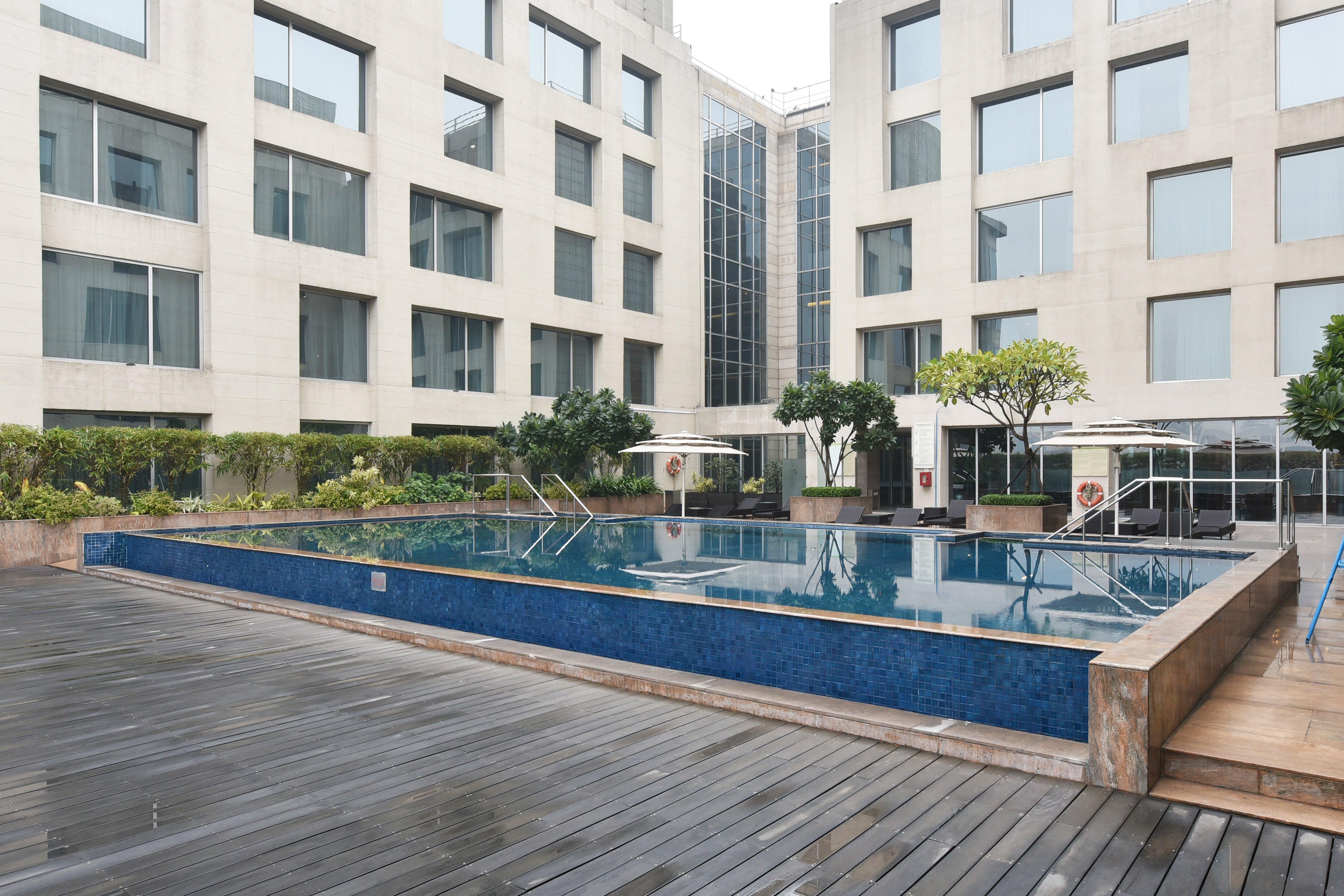 Holiday Inn New Delhi Aerocity By Ihg Exteriér fotografie The photo shows a modern outdoor swimming pool situated in a courtyard surrounded by several buildings. The pool has a sleek design with a blue tiled edge and is bordered by wooden decking. There are a few trees and plants around the area, adding som