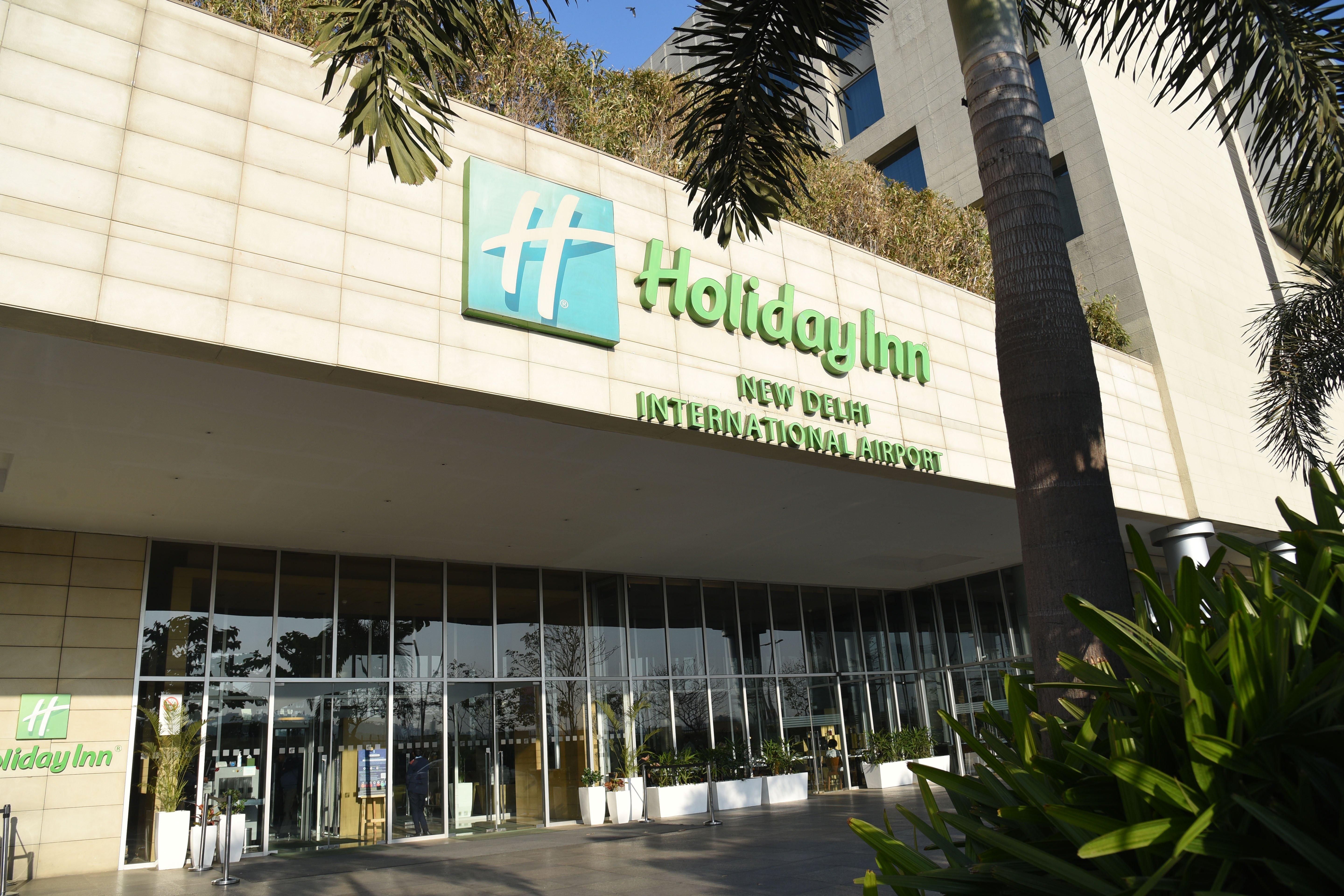 Holiday Inn New Delhi Aerocity By Ihg Exteriér fotografie The photo shows the exterior of a Holiday Inn hotel, specifically the Holiday Inn New Delhi International Airport. The building features a modern design with a prominent green and white sign displaying the hotel name. There are large glass doors at t