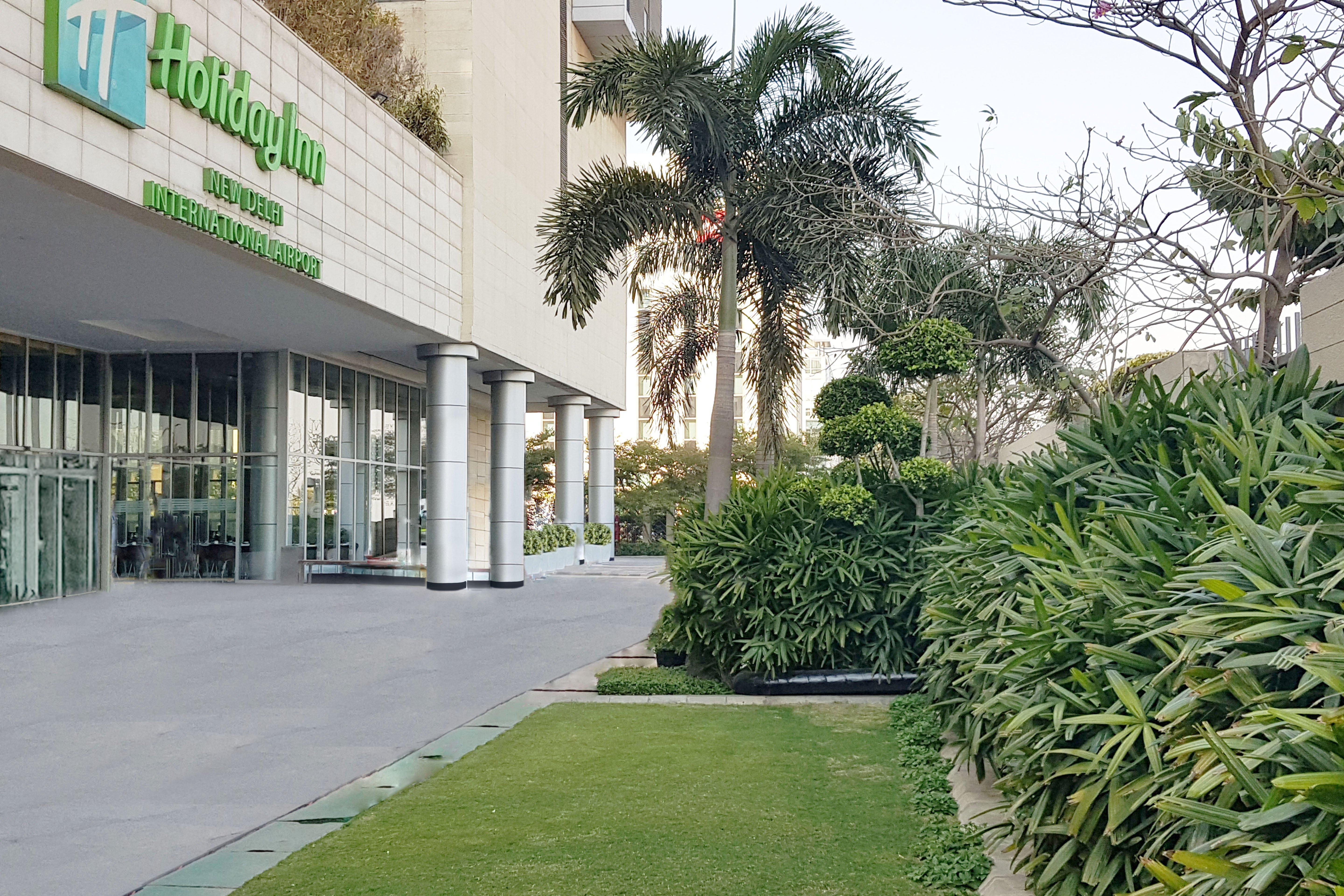 Holiday Inn New Delhi Aerocity By Ihg Exteriér fotografie The photo shows the exterior of a Holiday Inn hotel. In the foreground, there is a well-maintained green lawn and various ornamental plants, creating a pleasant garden area. The building features large glass doors and columns, and the hotel's name an